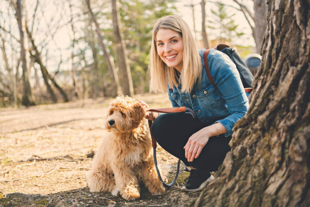 Woman Kneeling Beside A Mini Labradoodle Outdoors